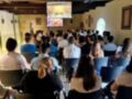 Una sala de conferencias con una pantalla grande en el centro, mostrando imágenes antiguas. La audiencia está sentada en filas de sillas coloridas, mirando hacia adelante. La decoración incluye molduras en el techo y cuadros en las paredes.