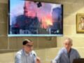Dos hombres sentados en una mesa con micrófonos, frente a un televisor que muestra imágenes de Notre-Dame en llamas.