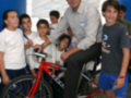 Un hombre en una bicicleta roja con niños alrededor, posiblemente en un evento o taller de ciclismo.