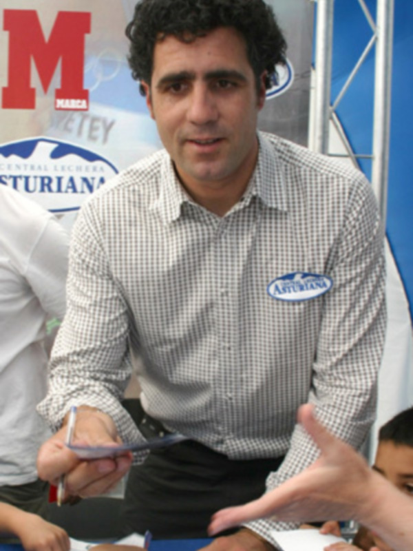 Un hombre en una camisa a cuadros firma un documento en un evento de marca. En el fondo, se puede ver una bandera con el nombre 'M' y 'Asturianas'.