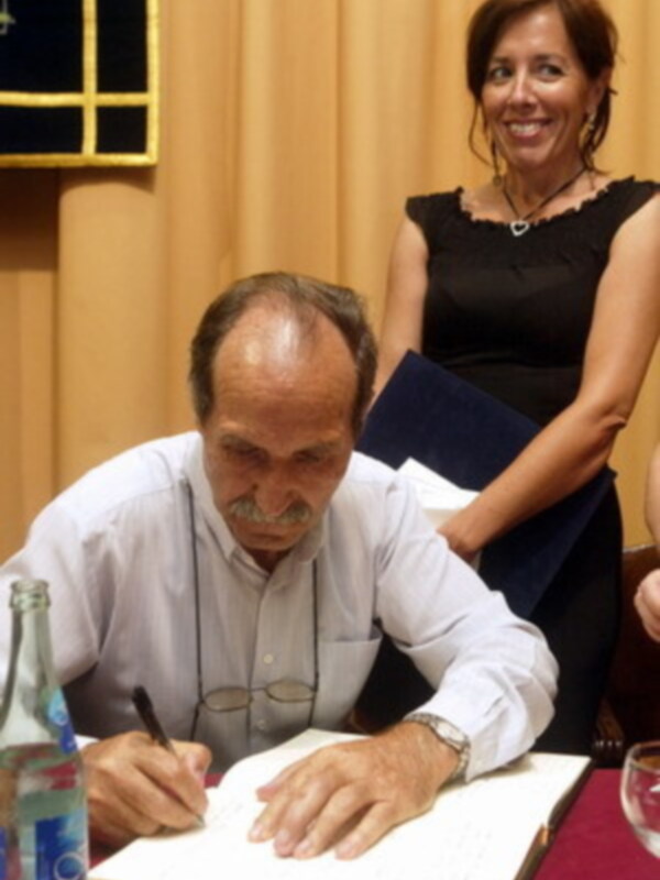 Un hombre mayor firmando un documento mientras una mujer observa en el fondo.