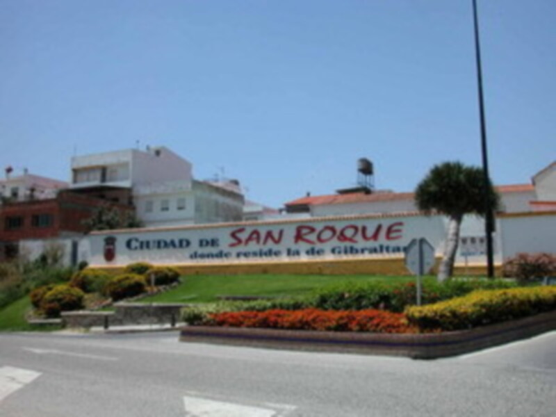 Un cartel de la ciudad de San Roque en una carretera, con edificios y plantas al fondo.