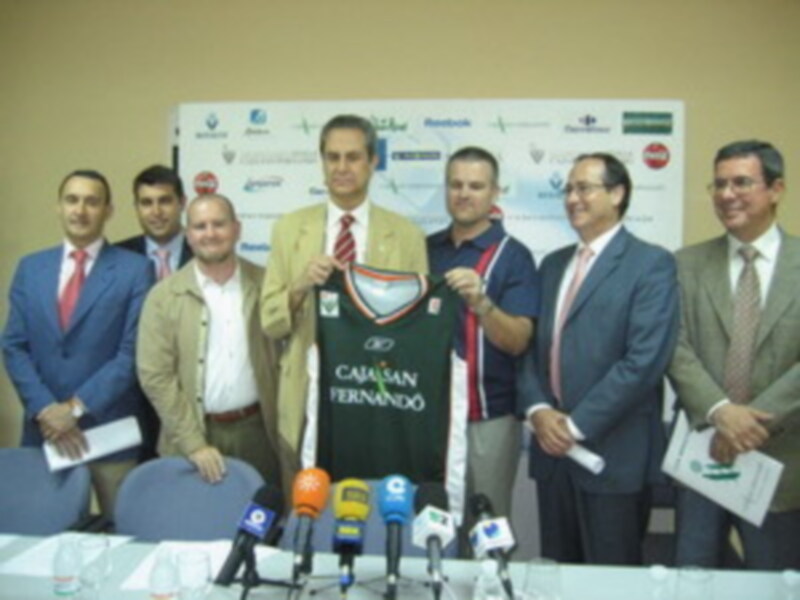 Presentación de una camiseta con el nombre "Fernando" en un evento formal con varios hombres en trajes y micros al frente.