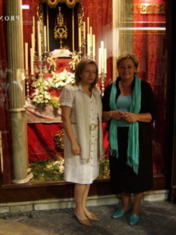 Dos mujeres posan frente a un altar con velas y flores, en un entorno religioso.
