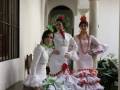 Tres mujeres vestidas con trajes de flamenca tradicional posan en un patio interior con arcos y columnas.