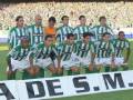 Equipo de fútbol posando para una foto en el campo, con uniformes verdes y blancos. El banner afuera dice "A DE S.M.".