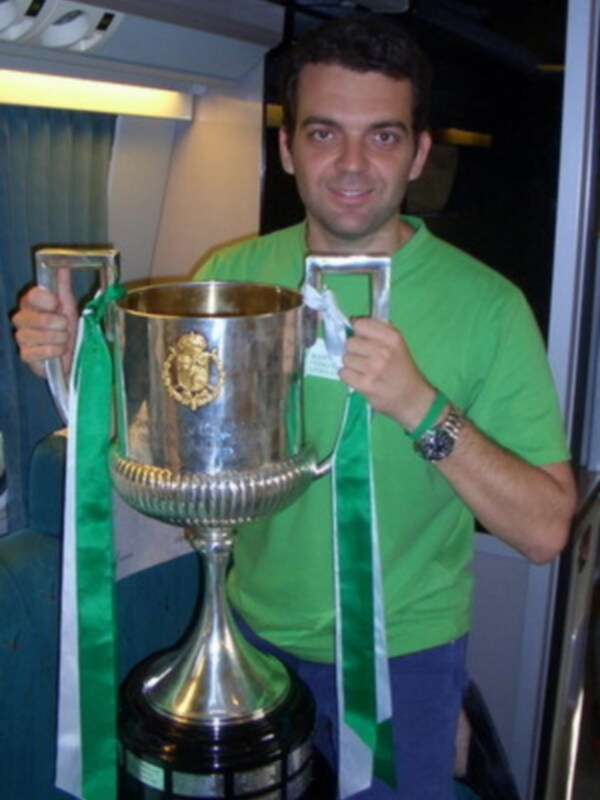 Un hombre sostiene una copa de plata con un emblema en la parte superior, vestido con una camiseta verde y pantalones azules. La imagen parece estar tomada en un vehículo, posiblemente un tren, con asientos y luces de techo visibles.