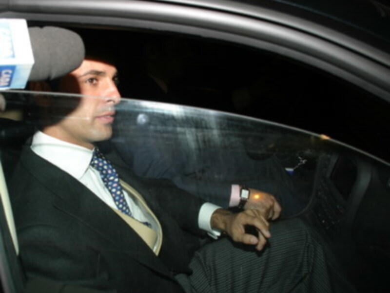 Hombre en traje y corbata sentado en el asiento trasero de un coche, con un micrófono de periodista en el lado del conductor.