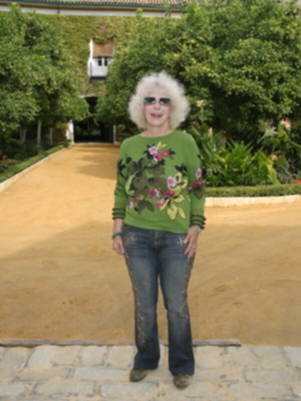 Persona con suéter verde bordado y jeans, posando en un patio con jardín y edificio tradicional en el fondo.