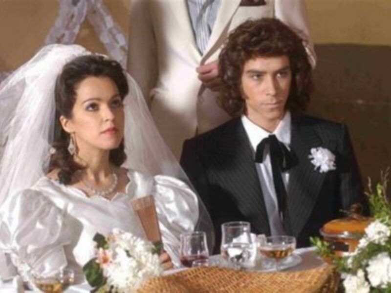 Una pareja en un banquete, el hombre está vestido de gala y la mujer lleva un vestido blanco y un velo, ambos sonríen y miran hacia la cámara.