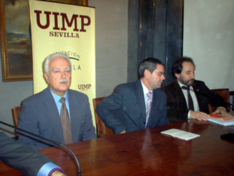 Conferencia en UIMP Sevilla, tres hombres en trajes sentados alrededor de una mesa con micrófonos y documentos.