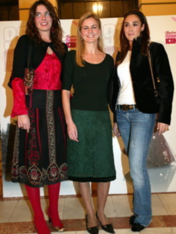 Tres mujeres posan juntas en un evento, vestidas con ropa formal y casual, en un ambiente interior con carteles de fondo.
