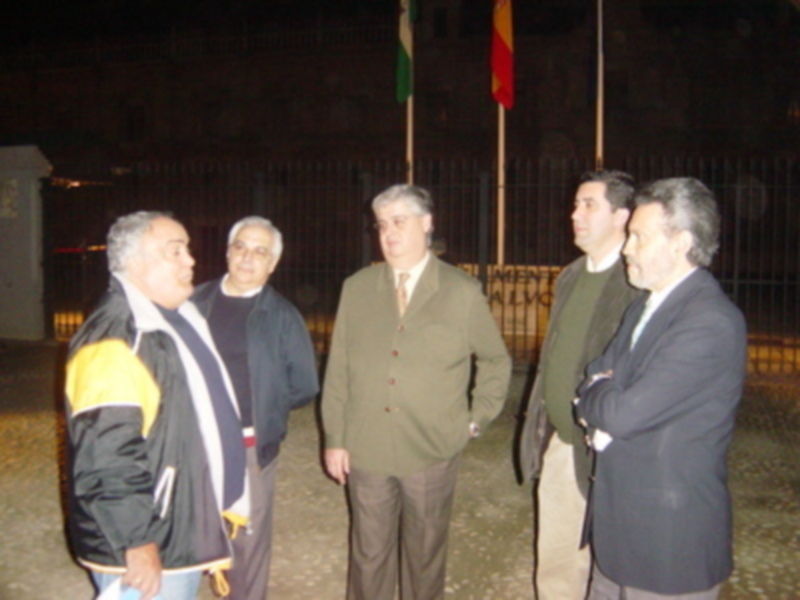 Cuatro hombres están de pie frente a una cerca con banderas en el fondo, posiblemente en una reunión o evento formal nocturno.