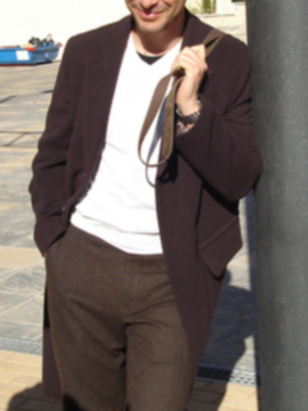 Persona sonriente en traje marrón y camisa blanca, posando al aire libre.