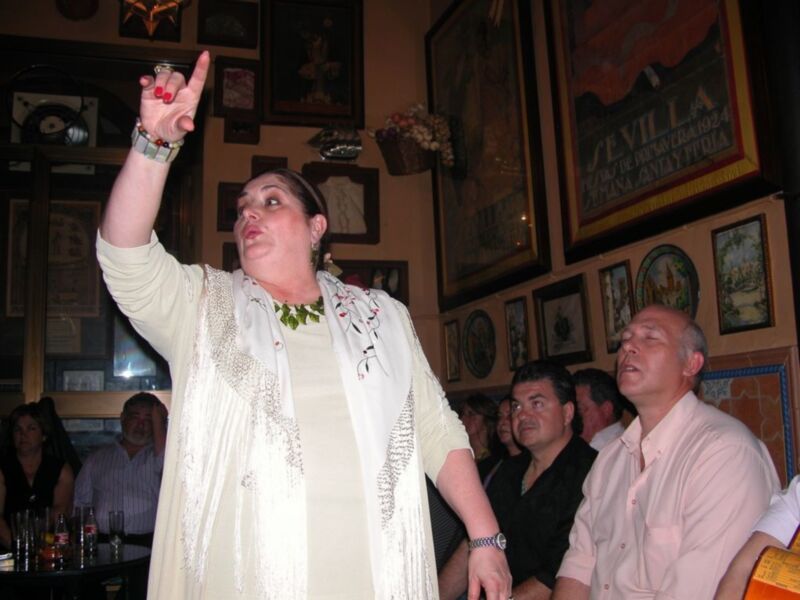 Una mujer en un bar levanta el brazo mientras otros observan. La pared está decorada con pinturas y carteles.