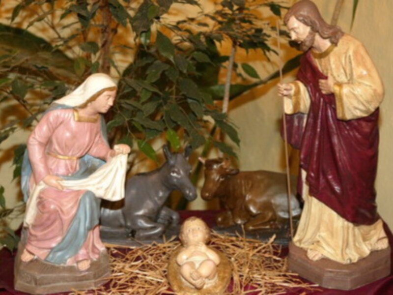 Escena de la Natividad con la Virgen María, el Niño Jesús y el Bebé Jesús en el pesebre, rodeados de animales como el burro y el reno, con un fondo de ramas verdes y un suelo de paja.