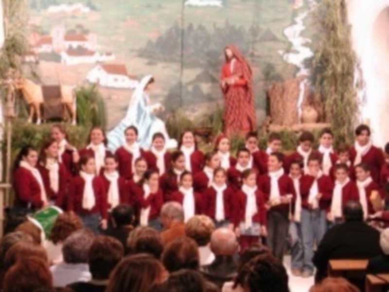 Concierto navideño con coro en trajes rojos y blancos, escenario con estatuas religiosas y decoración navideña.