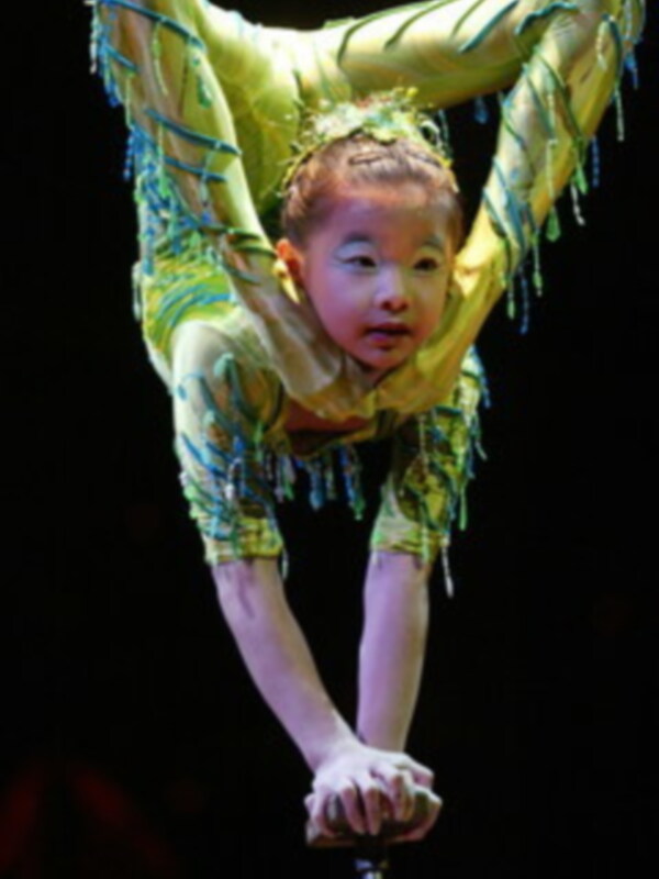 Niño en actitud de equilibrio sobre un poste, vestido con traje de circo con detalles de plumas, en escenario iluminado.