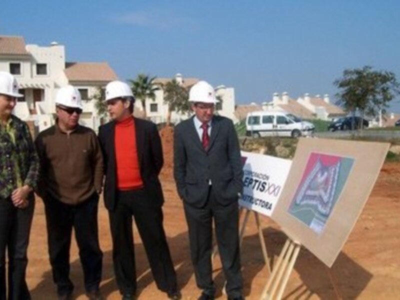 Equipo de construcción en un sitio de desarrollo inmobiliario, con paneles informativos y vehículos de trabajo al fondo.
