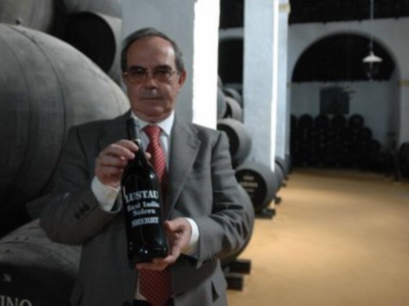 Hombre en traje sosteniendo una botella de vino Sherries en una bodega con barricas de madera al fondo.