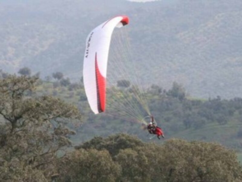 Paracaidista desciende con su paracaídas en un paisaje montañoso y boscoso.