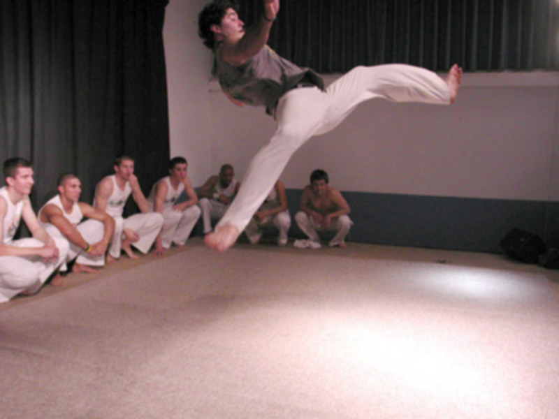 Un hombre realiza un salto en el aire, mientras que otros seis observan desde la distancia. El fondo es una cortina de color claro y el suelo es beige.