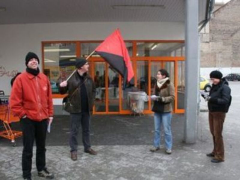 Grupos de personas en una plaza, algunas con banderas rojas y negras, posiblemente en una protesta o manifestación.