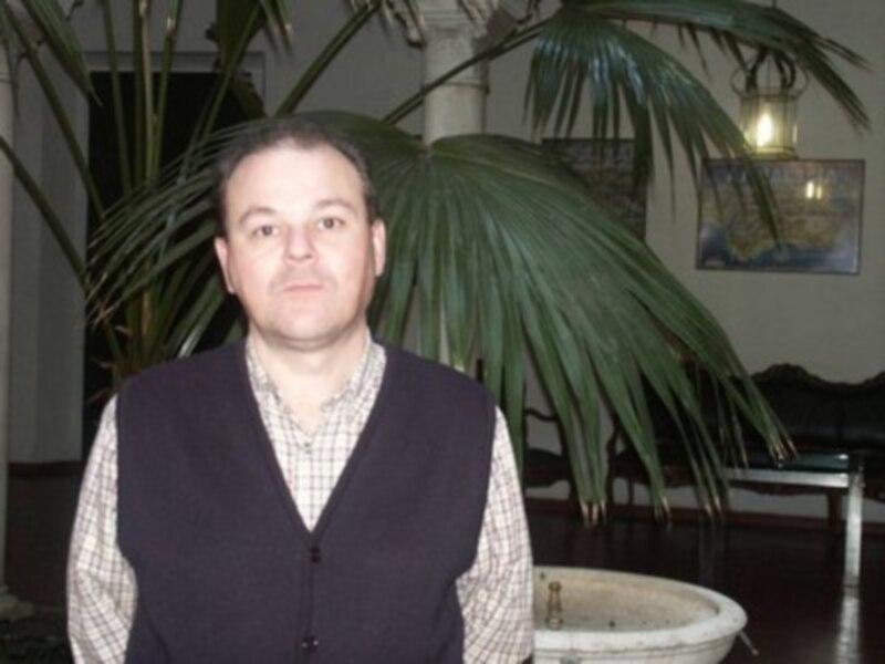 Hombre en chaleco y camisa a cuadros, fondo con planta y lámparas