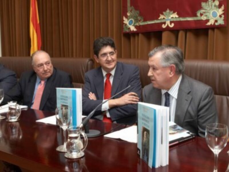 Tres hombres en trajes discutiendo en una sala formal con libros y vasos de agua en la mesa.