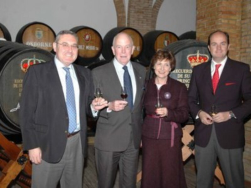 Celebración en bodega con barricas de vino y personas con copas de vino.