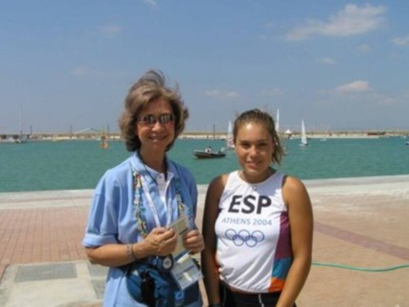 Dos personas posan frente al mar, con una camiseta de 'ESP' y medallas olímpicas, con el fondo de barcos y un cielo azul claro.