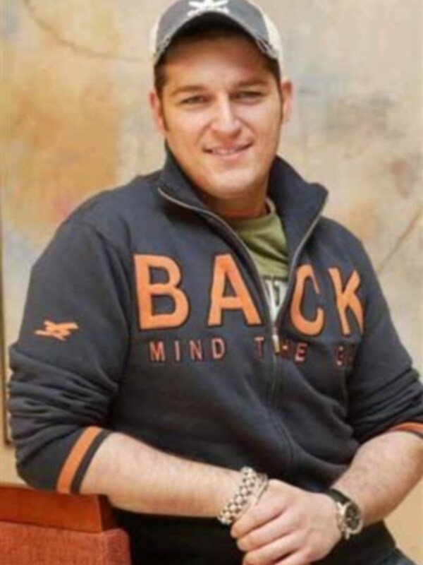 Un hombre con una sudadera negra con letras en rojo y una gorra de béisbol, posando con una sonrisa en el fondo de una pared de ladrillo.