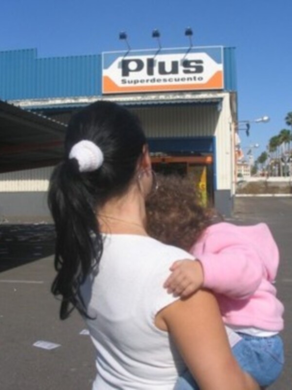 Mujer sosteniendo a un bebé frente a un supermercado 'Plus Superdescuento'.