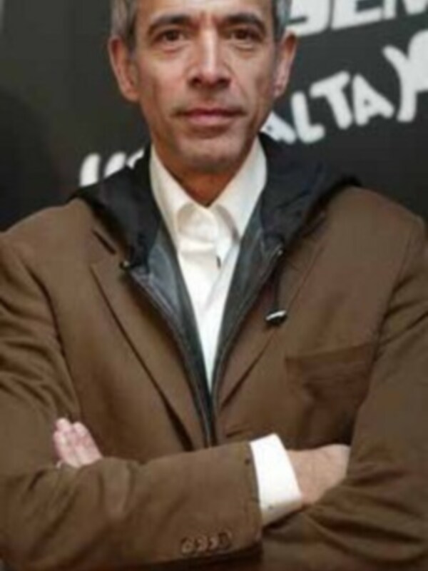 Un hombre con cabello gris y barba, posando con los brazos cruzados. Lleva una chaqueta marrón y una camisa blanca. La imagen parece ser de un evento o presentación, con letras en el fondo que no son legibles.