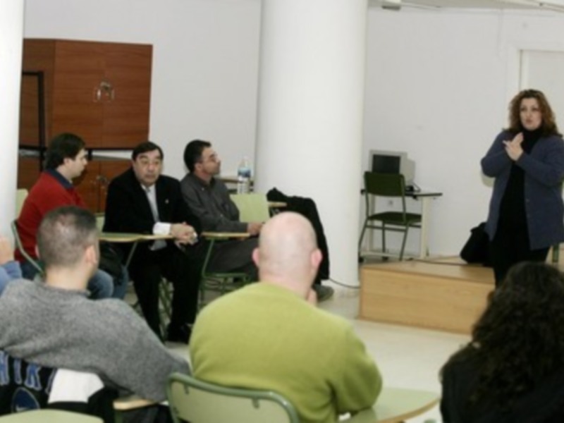 Una persona hablando en una sala con otros asistentes sentados en sillas. El ambiente parece ser un espacio de reuniones o conferencias.
