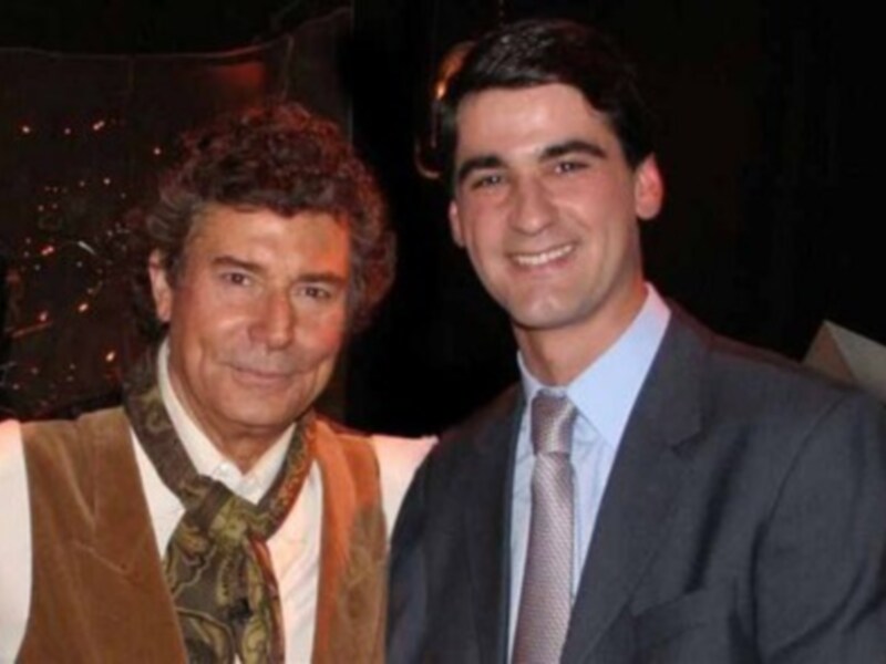 Un hombre en traje azul y corbata blanca posa junto a un personaje de la televisión en una escena de una película.