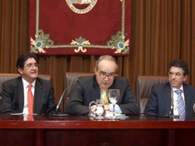 Tres hombres en trajes sentados en una mesa con micrófonos y copas de vino, en un ambiente formal con cortinas y un escudo en la pared.