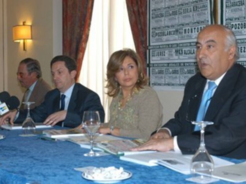 Tres personas sentadas en una mesa, con un fondo que incluye carteles y una lámpara de pared.