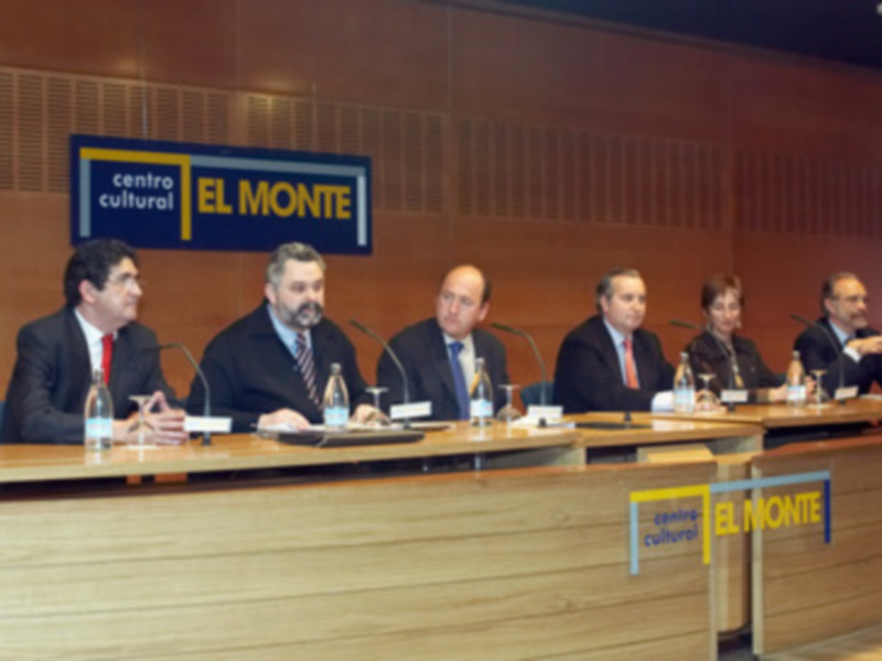 Una reunión formal en un centro cultural, con varios hombres sentados en una mesa, posiblemente discutiendo sobre un tema importante. La pared detrás de ellos tiene un cartel con el nombre 'El Monte'.