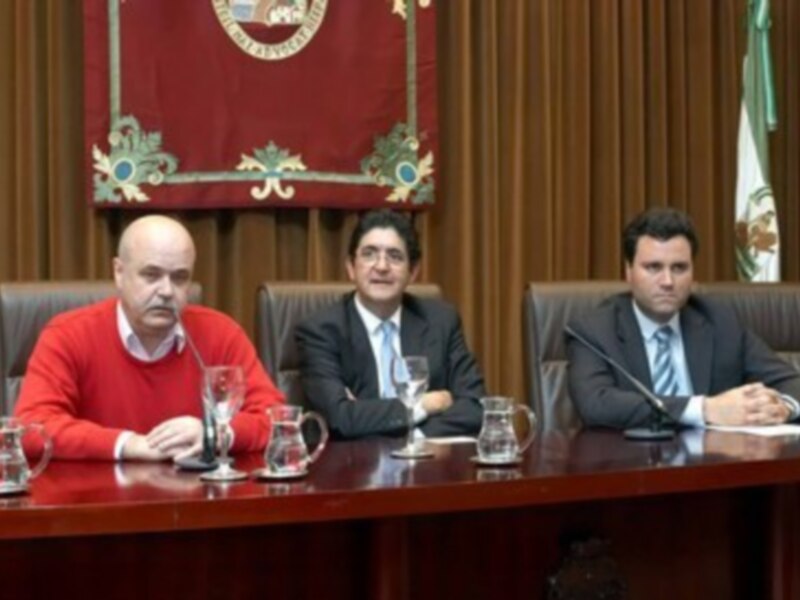 Reunión formal en una sala de conferencias con tres hombres sentados en una mesa, uno en traje rojo, otro en traje azul y otro en traje negro, con telón rojo y escudo en el fondo.