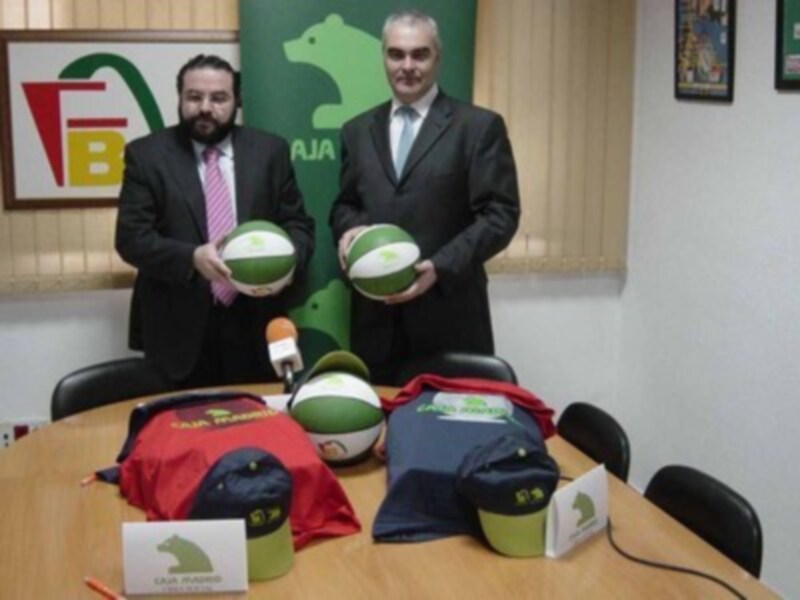 Dos hombres en trajes sostienen pelotas de baloncesto en una sala con equipamiento deportivo y logos en el fondo.