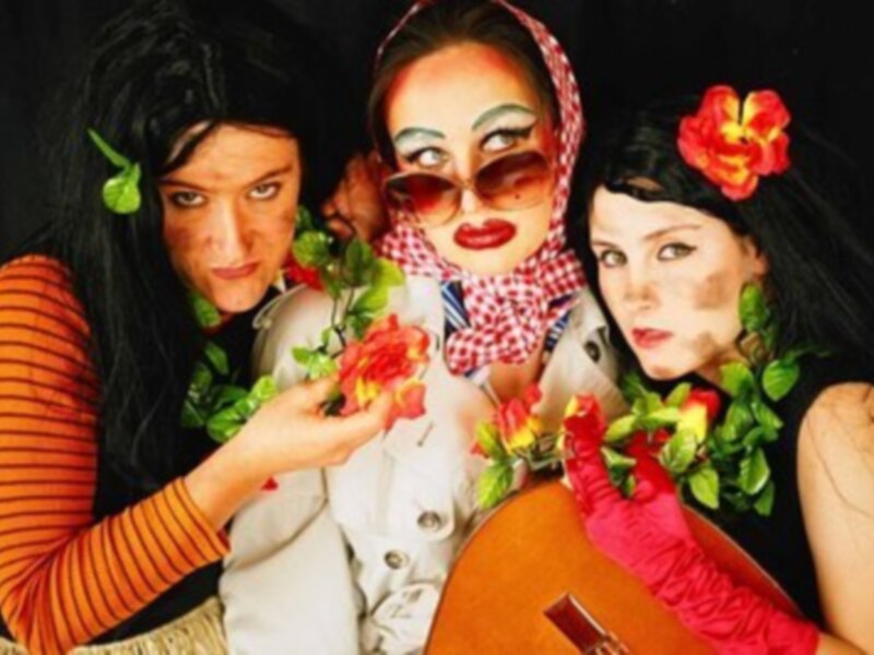 Tres mujeres en trajes coloridos y accesorios florales posan juntas con expresiones divertidas.