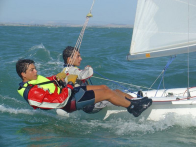 Dos personas disfrutan de la vela en el mar, uno está sentado en una tabla mientras que el otro controla las cuerdas. La imagen captura la emoción y la libertad del deporte de vela en el agua azul.