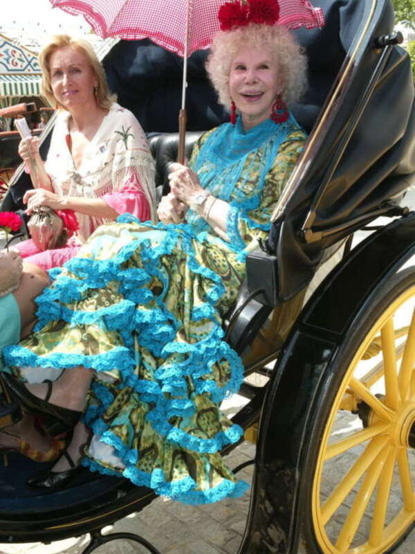 Dos mujeres en trajes tradicionales, una sosteniendo un paraguas con flores rojas, sentadas en un carro de caballos amarillo.