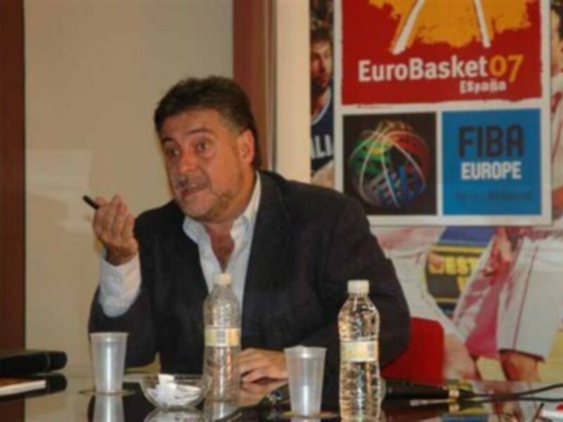 Un hombre en una conferencia de prensa, con una bandera de España y un cartel de EuroBasket 2007 en el fondo.
