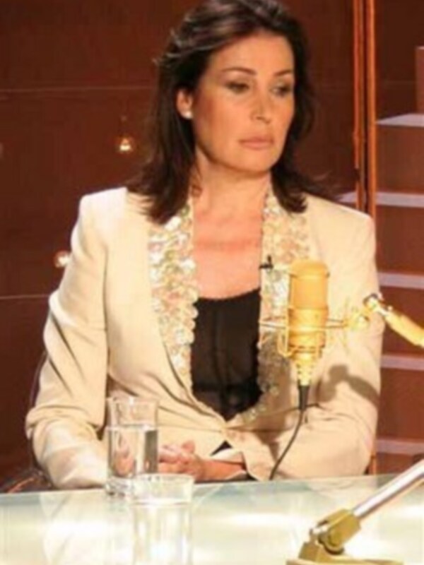 Una mujer en un entorno profesional, posiblemente en una entrevista o conferencia, con un microfono en frente de ella y una copa de agua en la mesa. La escena sugiere una transmisión de radio o televisión.