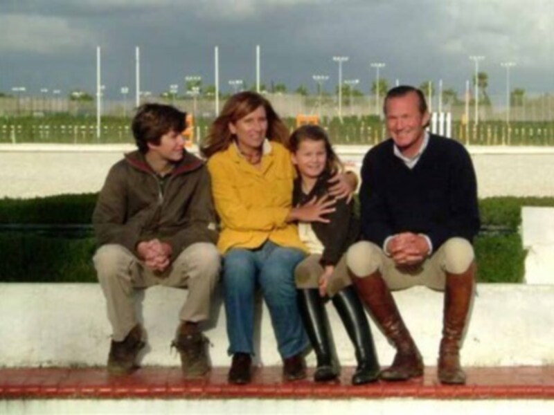 Una familia de cuatro personas sentadas en una banqueta, con un hombre y una mujer en el centro, y dos niños a sus lados. La escena parece ser en un parque o área pública, con palmeras y un cielo nublado en el fondo.
