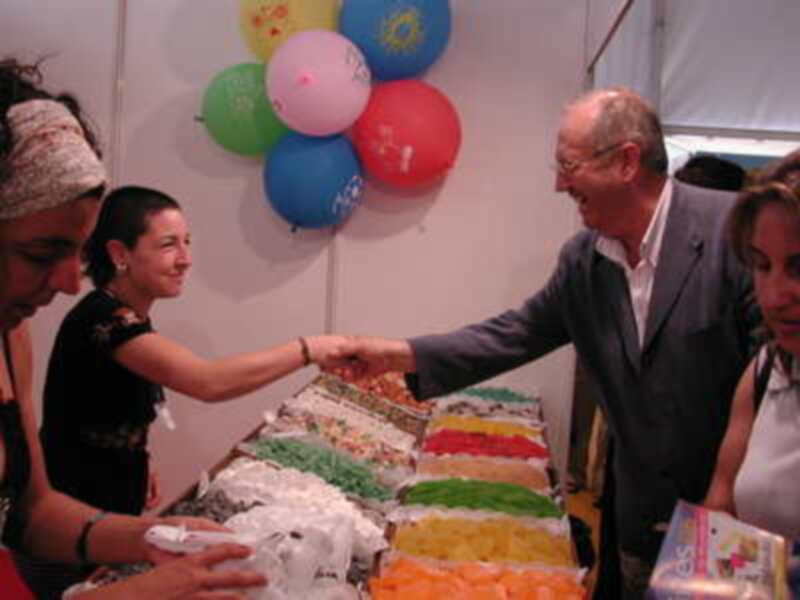 Persona saludando a otro en un mercado de dulces coloridos, con globos de colores al fondo.