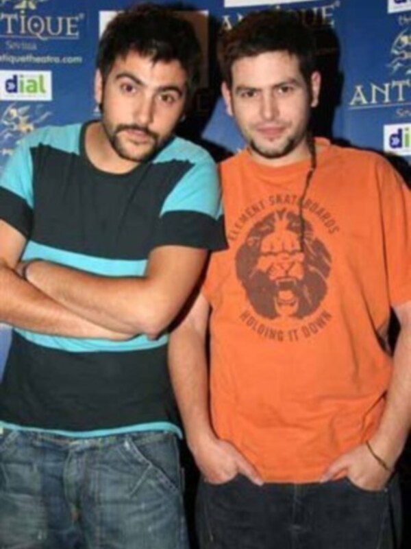 Dos hombres posan juntos en un evento, uno con camiseta azul y el otro con camiseta naranja, ambos con expresiones serias.