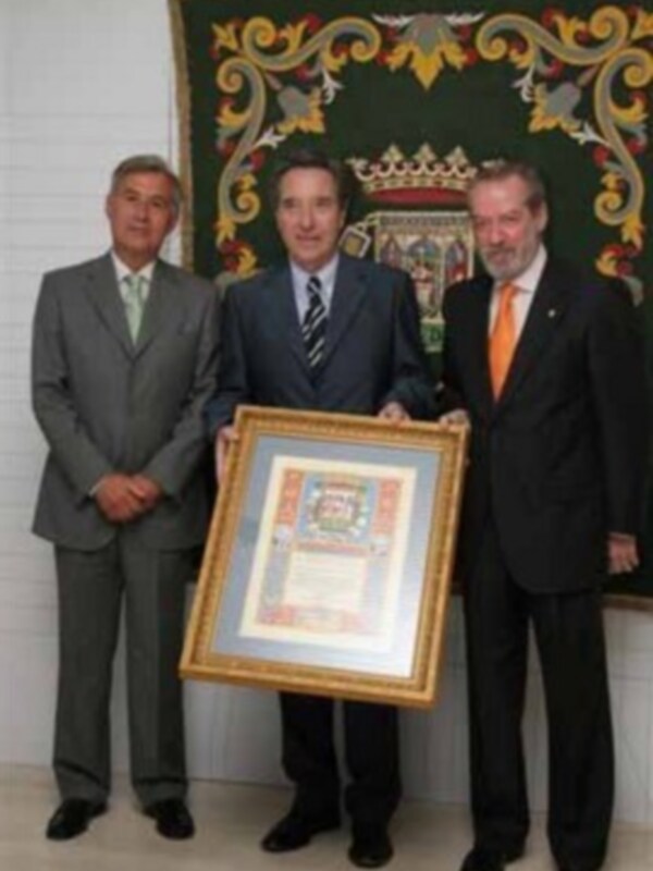 Tres hombres en trajes sostienen un marco con un documento, posando frente a un tapiz decorativo.